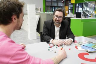 Portrait de FibreTigre installé à une table avec des dés. Il est à droite de l'image, assis face à un homme de profil à gauche.