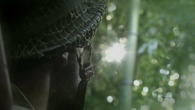 En très gros plan, la tête d'un soldat de dos. Face à lui, le soleil perce à travers le feuillage des arbres.