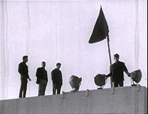 Des ouvriers debout sur un toit. Un drapeau flotte au vent.