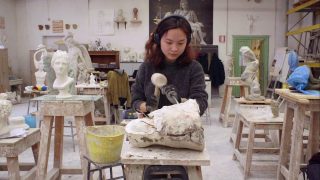 Dans un atelier, une jeune femme sculpte un bloc de pierre avec un marteau.
