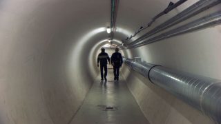 Dans un tunnel arrondi, deux hommes en combinaison de travail marchent.
