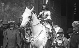 Photographie en noir et blanc d'une jeune femme costumée en Jeanne d'Arc, en armure, sur un cheval blanc