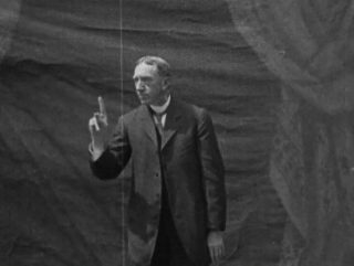 George Veditz, en costume devant une feuille de décor représentant un rideau de théâtre, signe face à la caméra.